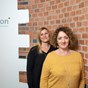 2 women standing in front of a wall, logo behind them 