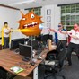 3 men dressed as superheros in office blasting light at an orange monster floating above a desk. 
