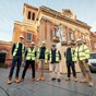 A town hall building in the background with six workmen wearing hi-vis yellow vests and hard hats are stood in front in town centre