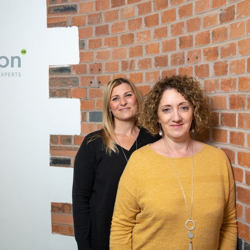 2 women standing in front of a wall, logo behind them