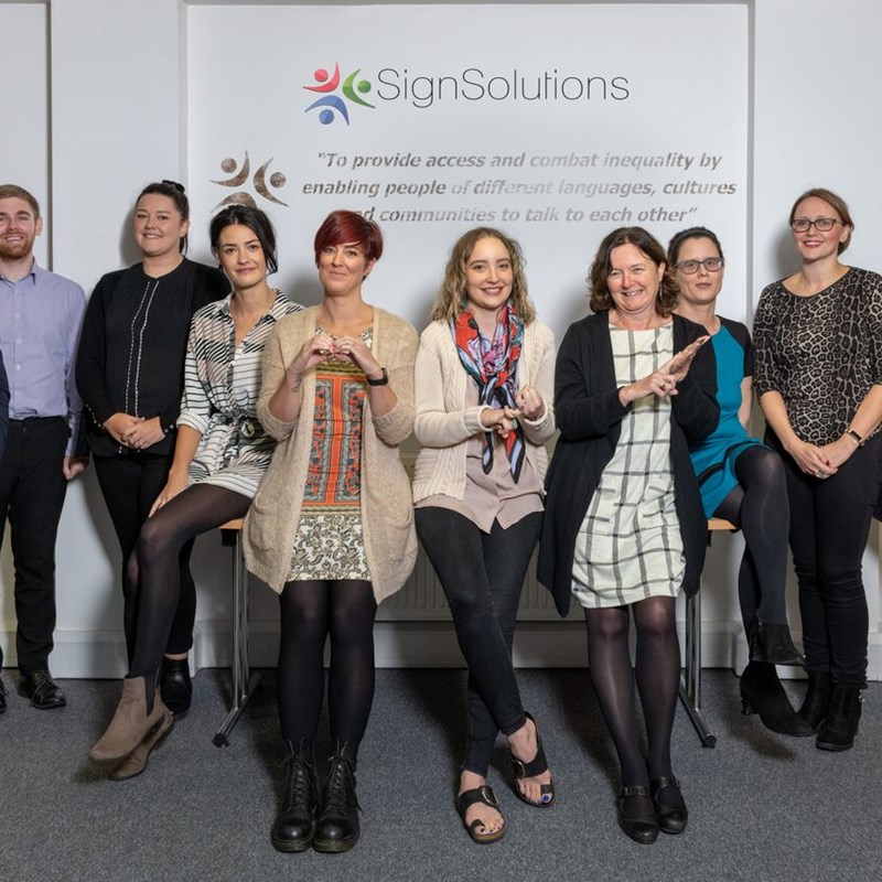 Group of people standing in front of company name 'Sign Solutions', the three at the front are signing, one the letter B, the second the letter S the third the letter L